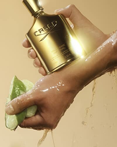 Hand holding a golden perfume bottle with the brand name Creed, next to a piece of fruit.